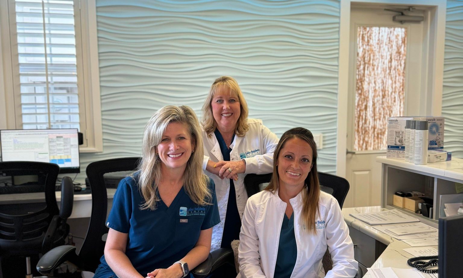 Image shows three smiling women at the front desk of Lake Baldwin Dental, who are dedicated to making LBD the best dentist in Orlando!
